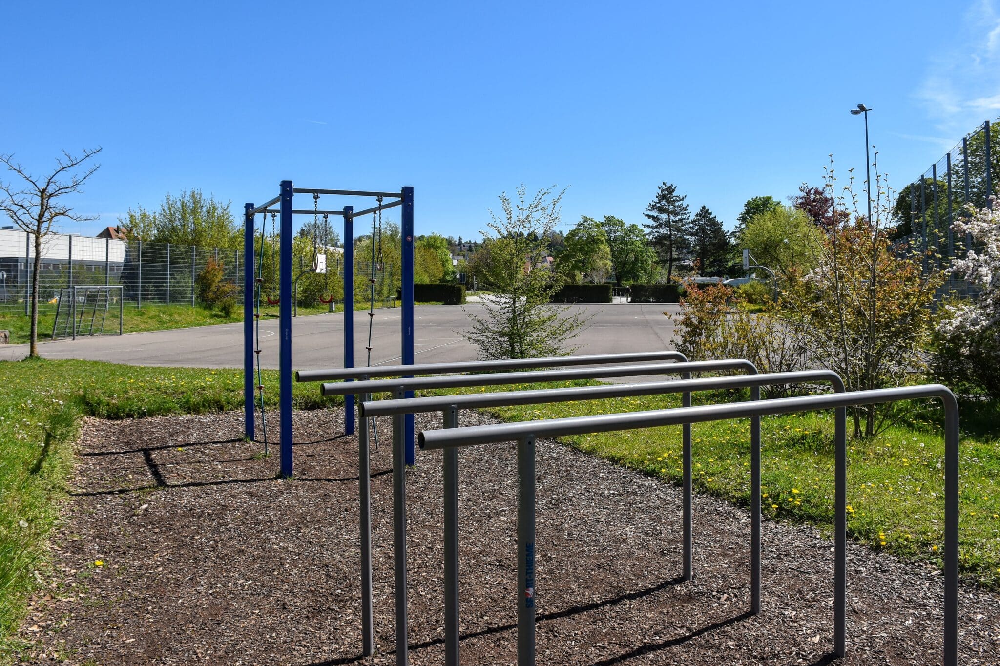 Calisthenics-Anlage mit vier niedrigen Reckstangen und einer blauen Fitnessstation mit vier Calisthenics-Ringen und zwei Kletterseilen. Im Hintergrund sind der Multifunktionsplatz mit Basketballkörben und Fußballtor sowie Bäume und Sträucher zu sehen.